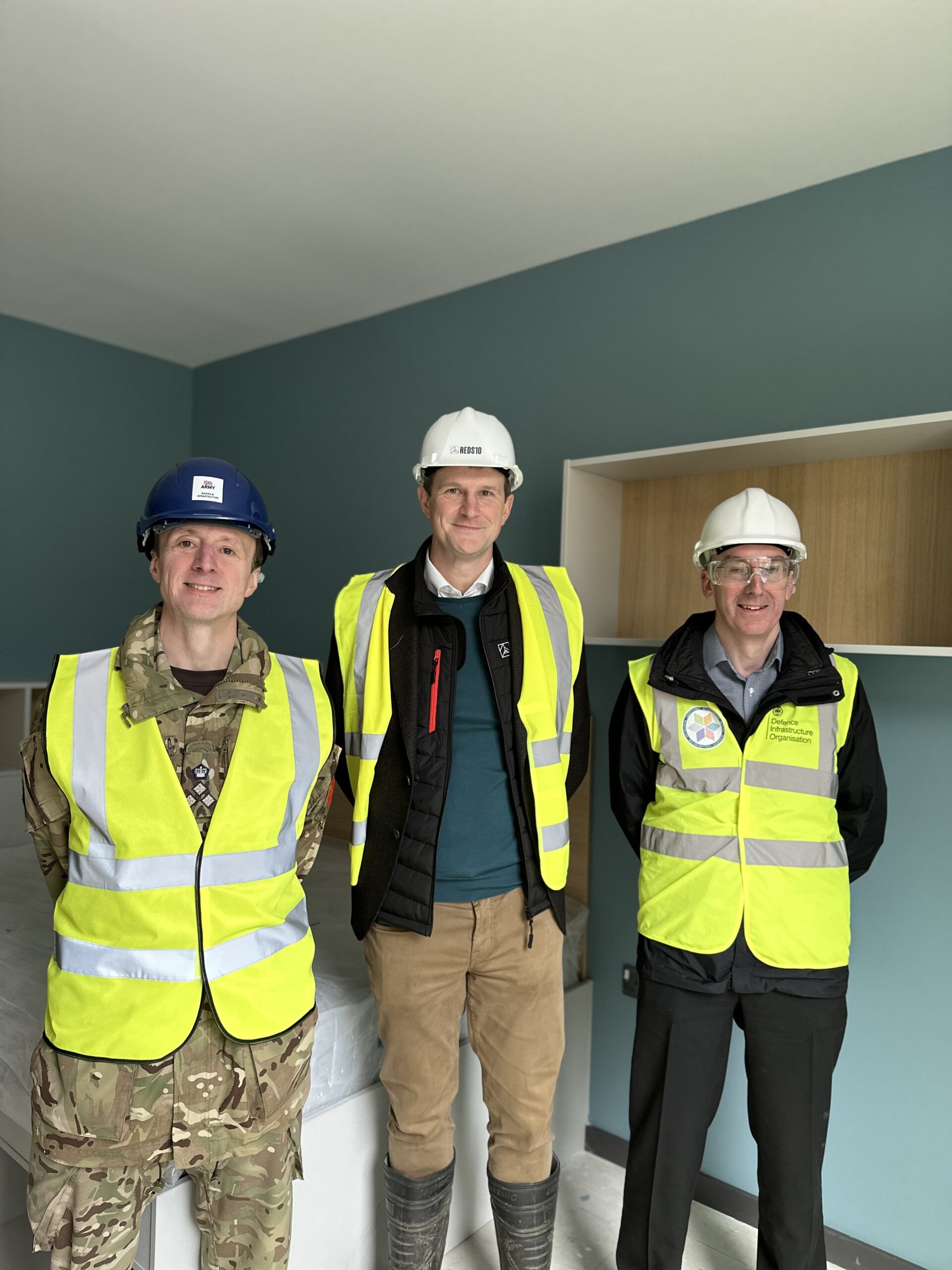 a photo of 3 men standing in a row inside a blue-painted bedroom. They are wearing construction vests and hard hats: one is in Army uniform. There is an empty shelving unit on the wall behind them.