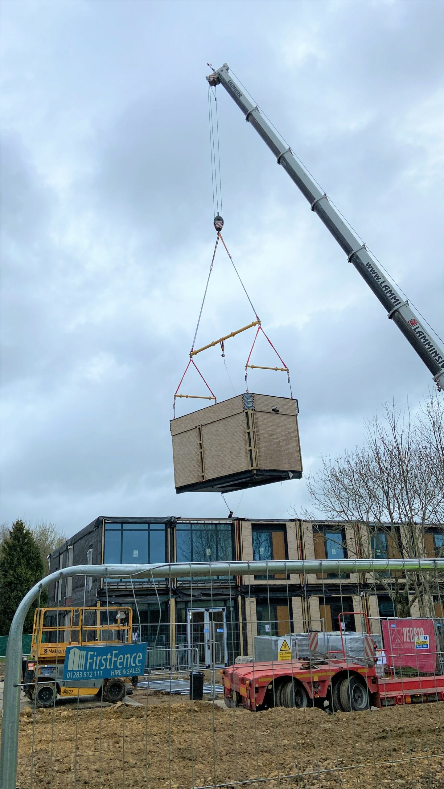 a photo of a crane lifting a large rectangular module onto a 2-storey building under construction. Construction machinery and trees are visible in the surroundings.