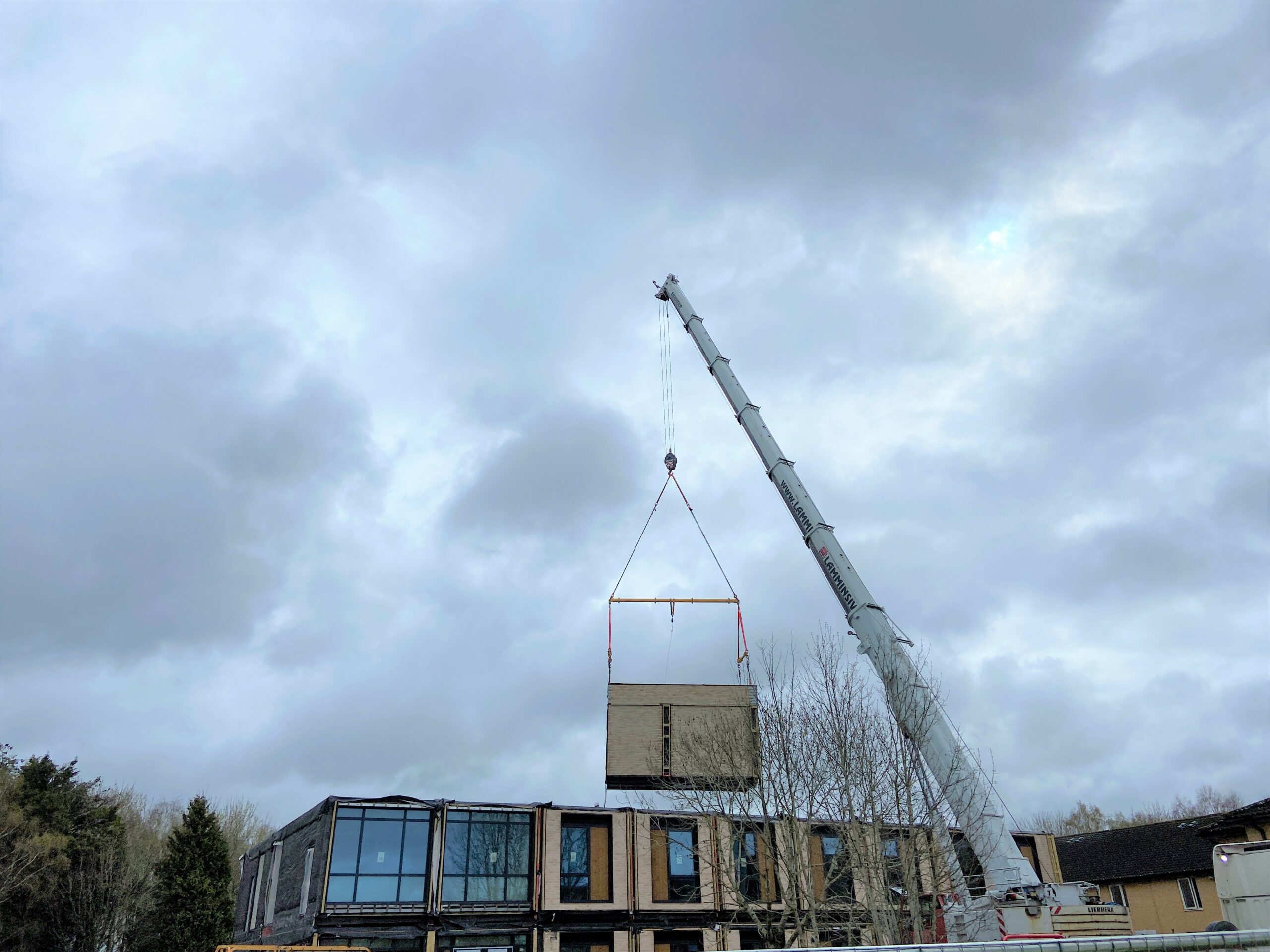 a photo of a crane lifting a large rectangular module onto a 2-storey building under construction. Construction machinery and trees are visible in the surroundings.