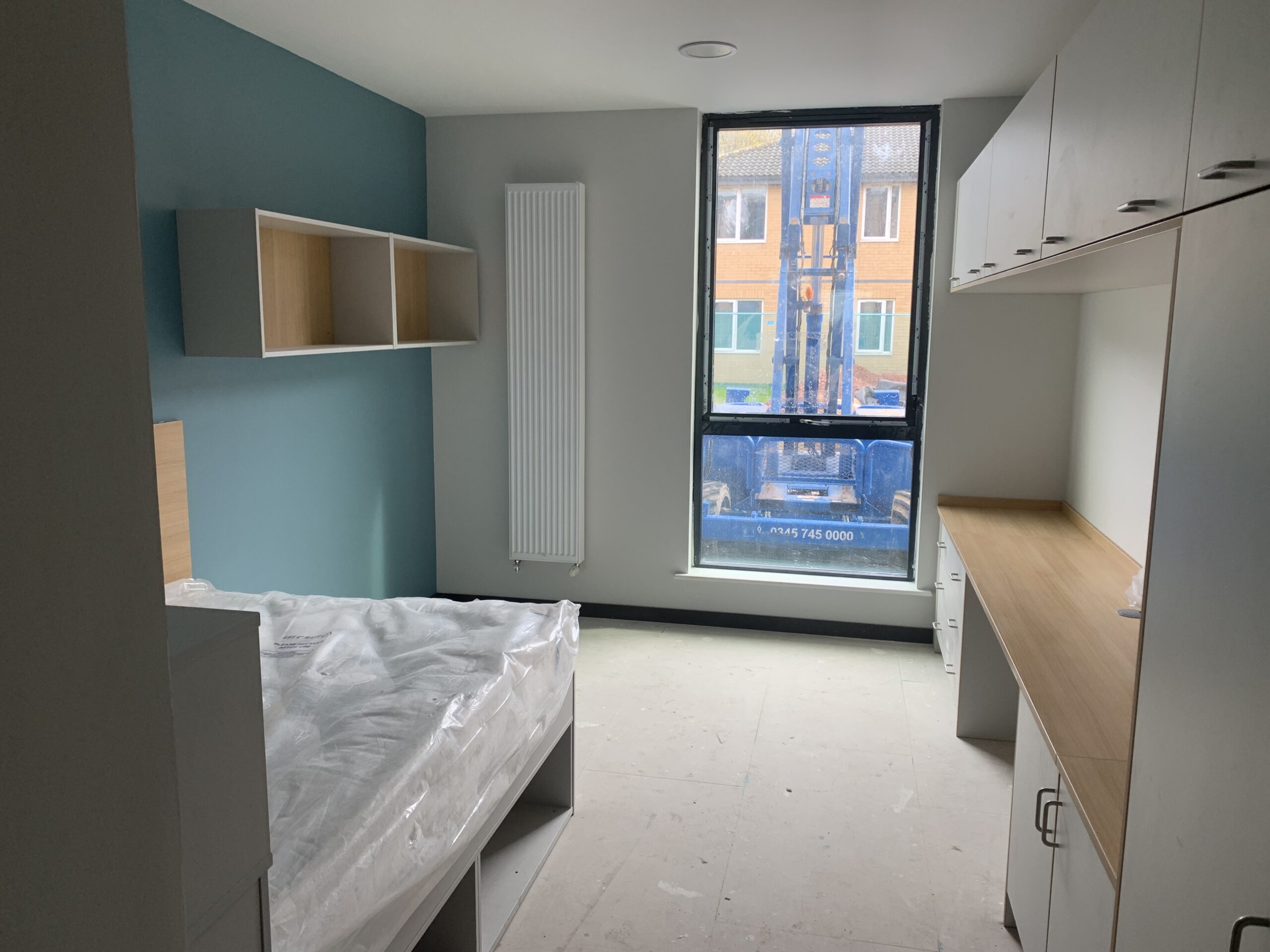 a photo of an empty bedroom with a bed on the left and built-in cupboards with a desk on the right. There is a shelving unit on the wall above the bed and a wall-mounted radiator opposite the bed. There is a floor-to-ceiling window through which construction machinery is visible.